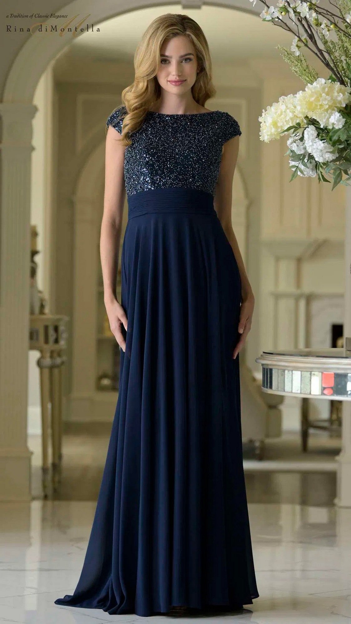 A woman poses in a well-lit, elegant room wearing the Rina Di Montella RD3419 long formal dress in navy, featuring a beaded bodice and flowy chiffon skirt.